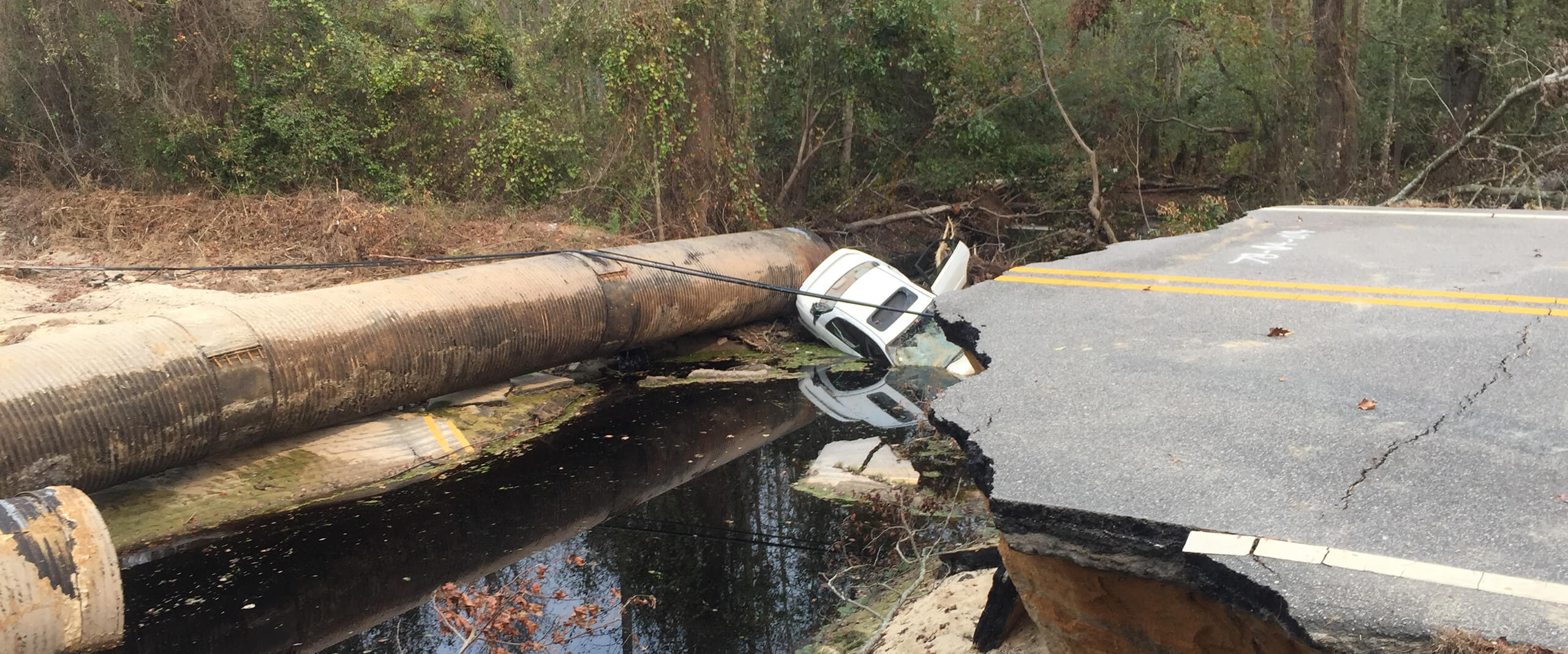 An abandoned vehicle is stuck in a ditch near a broken paved roadway after Hurricane Matthew's damage