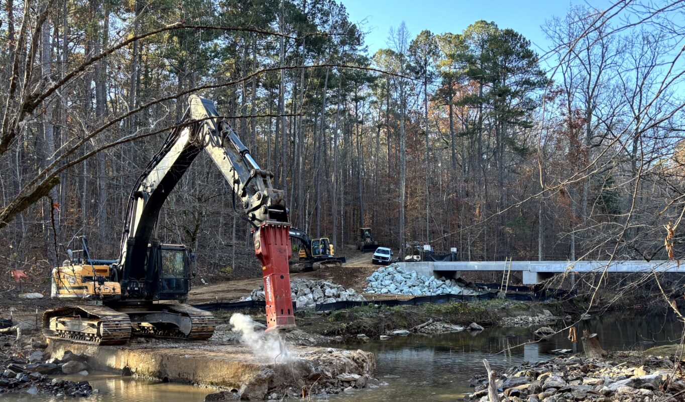 Excavator removing the old low-water crossing as its replacement bridge stands completed behind it